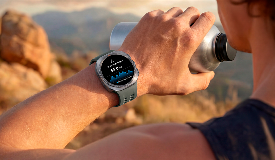 Um homem em uma trilha de montanha com terreno rochoso segura um copo de água enquanto usa um Galaxy Watch Ultra2 em Titânio Prata com Pulseira Marine Verde. O relógio exibe um trajeto de trilha com distância, ajudando na navegação em condições difíceis durante corrida em trilhas e escalada.