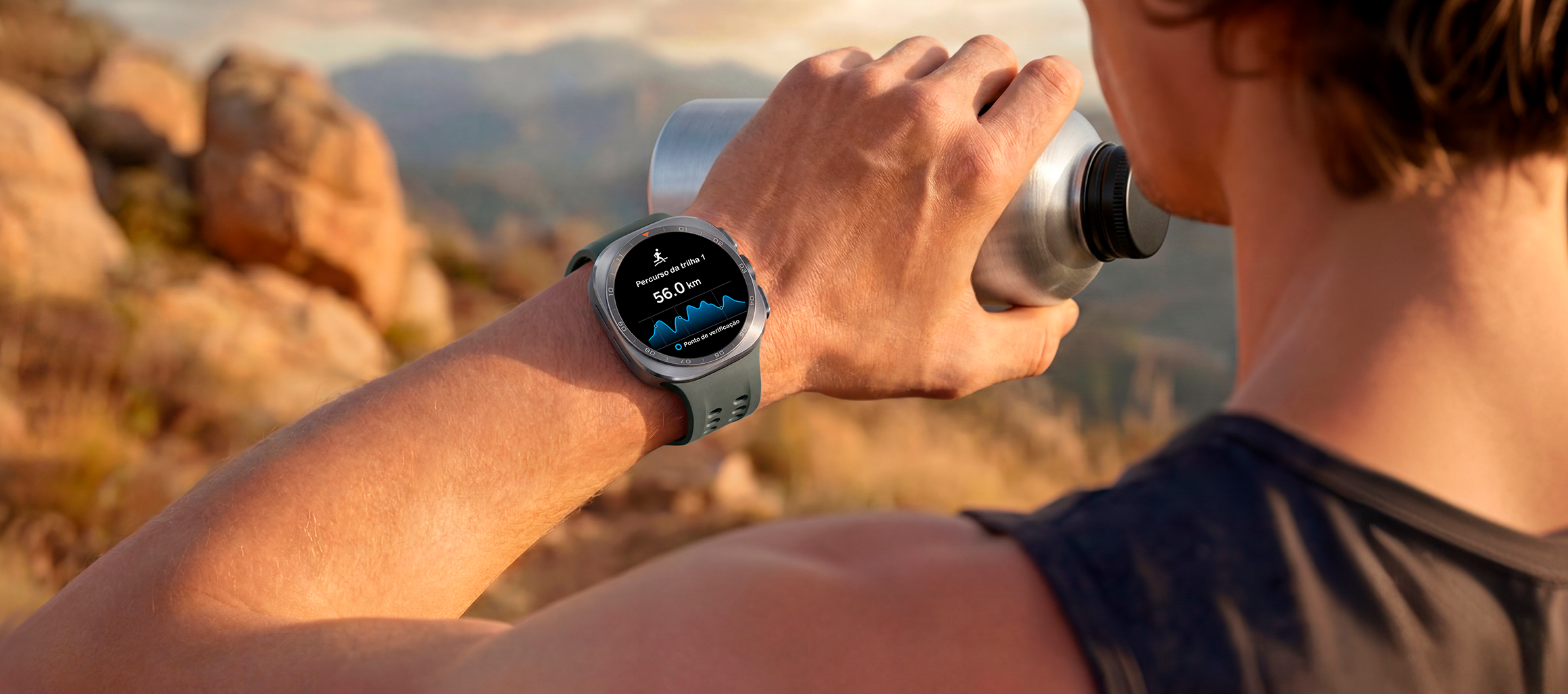 Um homem em uma trilha de montanha com terreno rochoso segura um copo de água enquanto usa um Galaxy Watch Ultra2 em Titânio Prata com Pulseira Marine Verde. O relógio exibe um trajeto de trilha com distância, ajudando na navegação em condições difíceis durante corrida em trilhas e escalada.