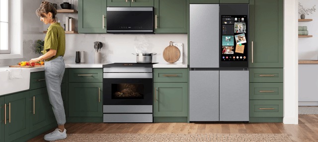 A modern kitchen setup featuring green cabinetry with a woman preparing food on the counter, a smart refrigerator with a display screen, and matching appliances.