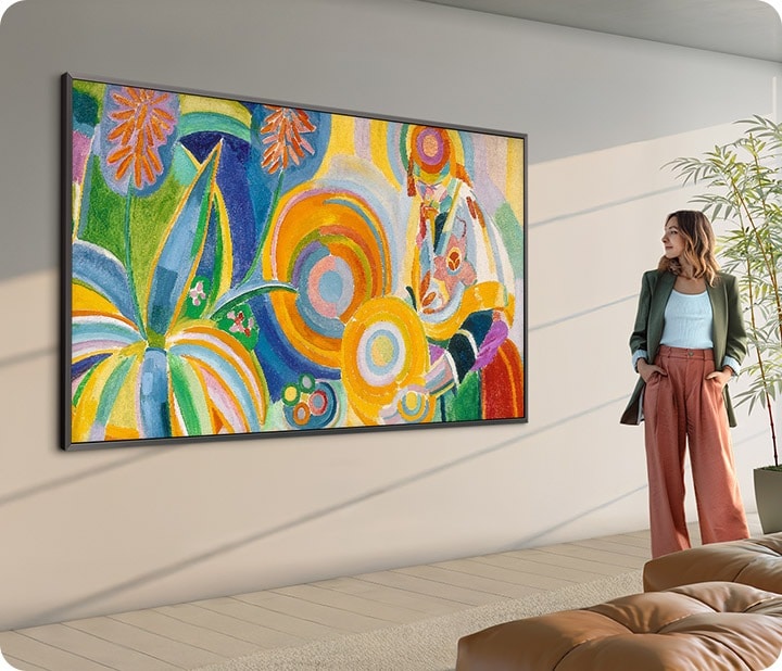 A large 98-inch The Frame TV displaying artwork in a bright modern living room, with a person standing nearby viewing the piece.