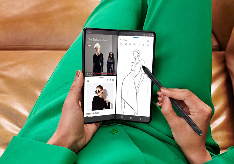 Hands holding unfolded Galaxy Z Fold3 5G and sketching with the S Pen Fold Edition. The Main Screen is in Multi Active Window mode. One half of the screen shows two instances of the Internet app open, one with a live stream of a fashion show and the other with a website showing images of the garments from the show. The other half of the screen is a sketch of a dress being done in Samsung Notes.