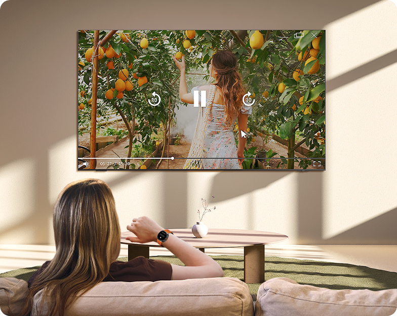 A woman controls a TV across the room with her Galaxy Watch. She gestures to press pause on a scene of a girl picking oranges.