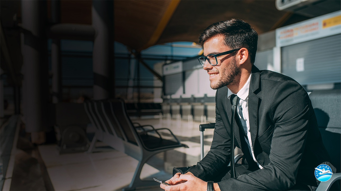 Молодой бизнесмен в очках смотрит перед ним, улыбаясь, сидя на станции с телефоном в руке. Логотип Air Miles показан в левом нижнем углу