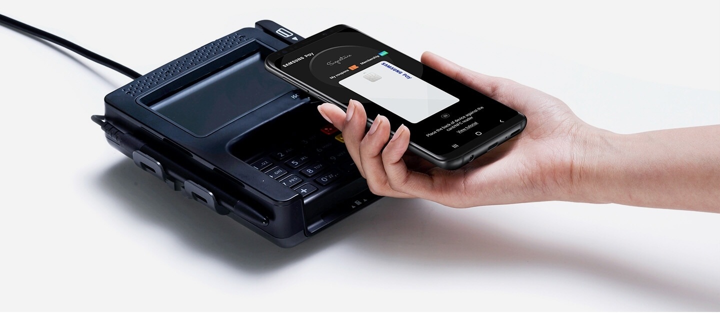 Imej tangan lurus memegang telefon pintar pintar dengan skrin di atas titik terminal jualan yang menunjukkan skrin dengan aplikasi Samsung Pay yang digunakan