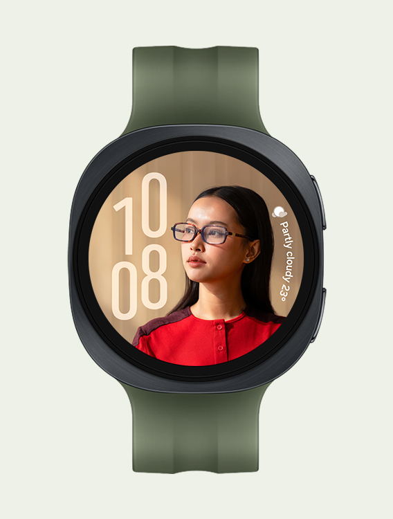 Vue de face et gros plan en angle de la montre Galaxy Watch9 équipée d'un bracelet de sport en olive, affichant un cadran personnalisé de type portrait avec l'heure à gauche et la météo à droite, sans chevauchement avec le visage de la personne.