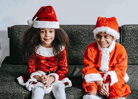 Bub und Mädchen in roter Weihnachtskostümen sitzen lächelnd auf der Couch.