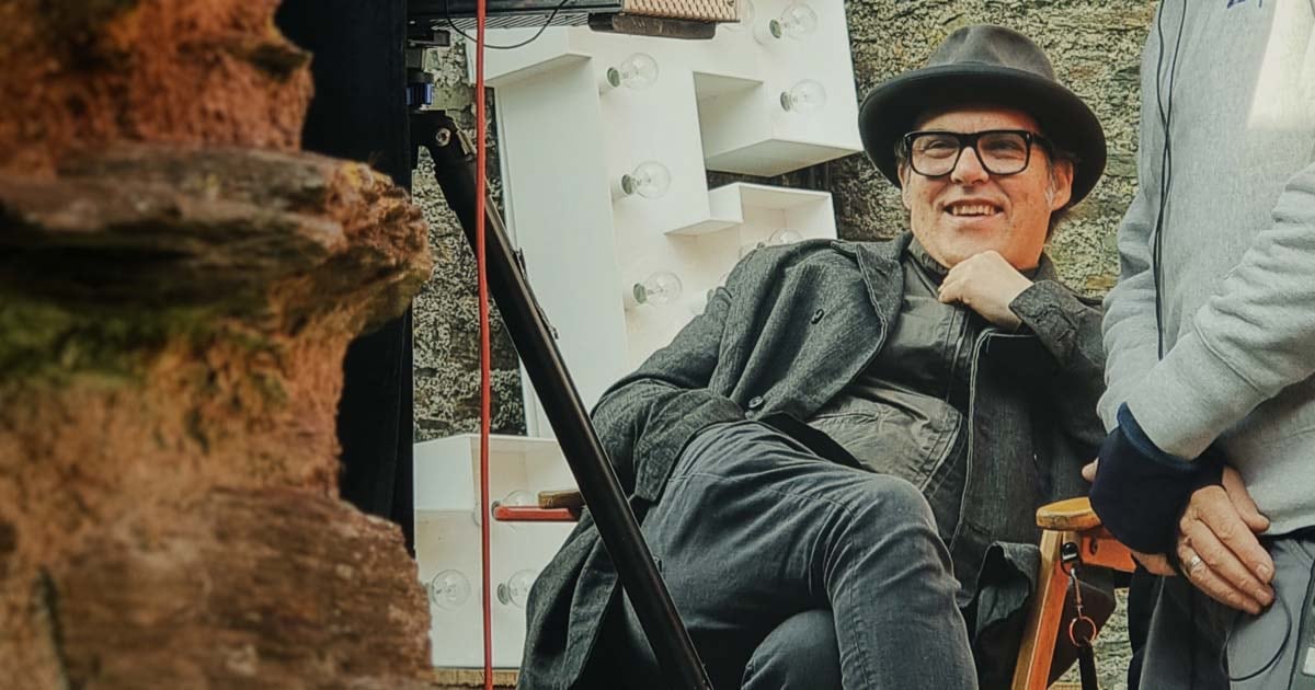 Joe Wright sitzt in einem Regiestuhl auf einem Produktionsset, schaut auf einen Monitor und lächelt.