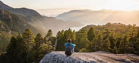 Rückansicht eines Mannes, der auf einem Felsen sitzt und auf eine friedliche Waldlandschaft mit einem See blickt.