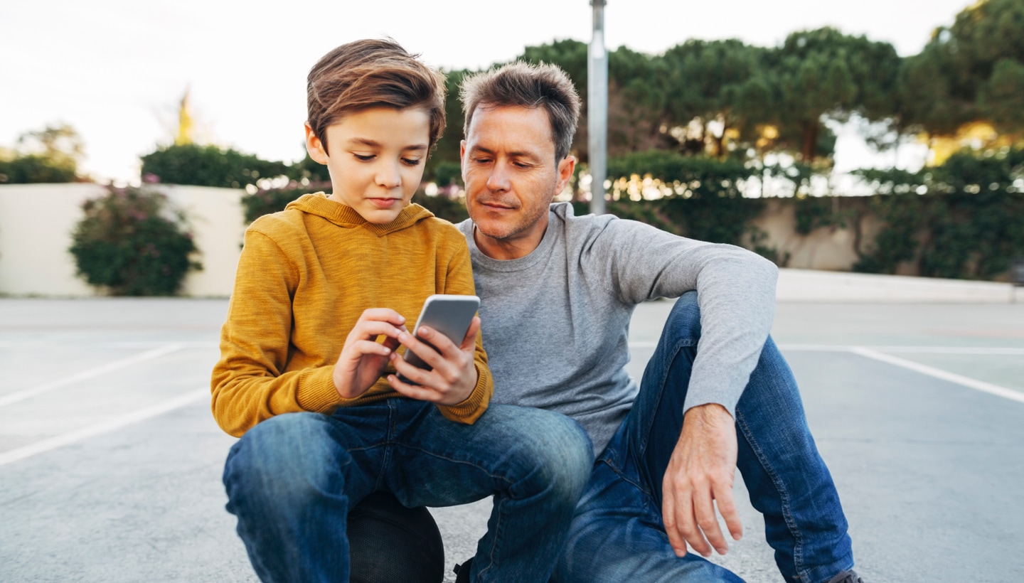 Ein im Freien aufgenommenes Foto eines Jungen, der auf einem Basketball sitzt und auf sein Smartphone sieht, während sein Vater neben ihm sitzt
