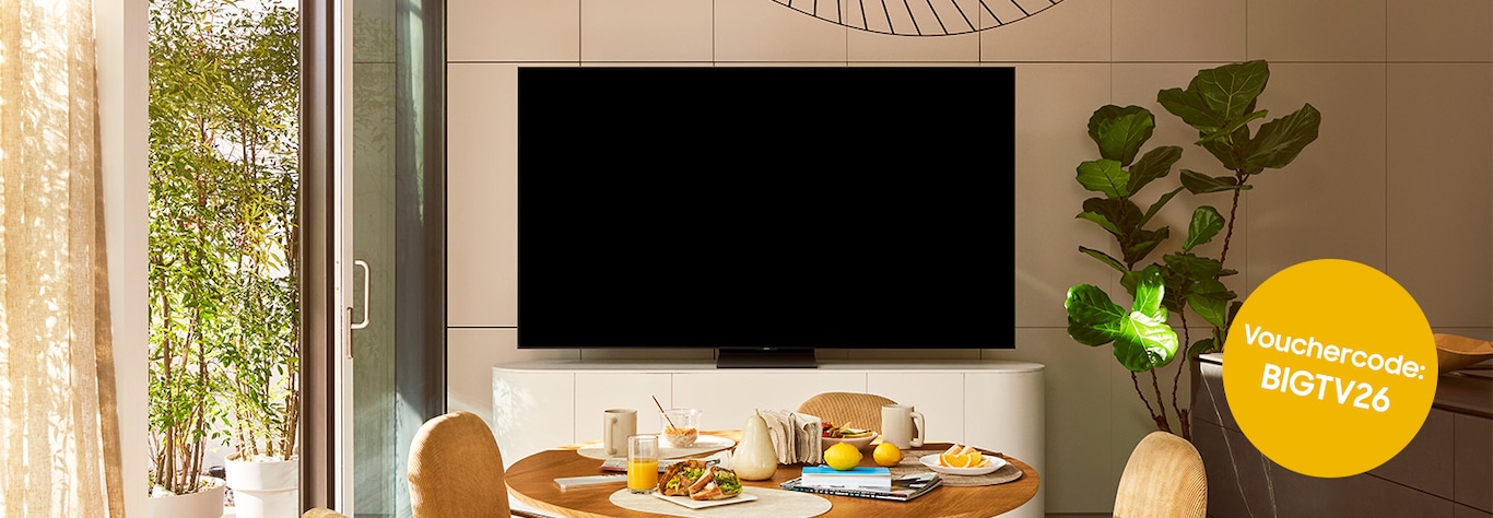 Ein gemütliches Wohnzimmer mit einem grossen Fernseher an der Wand, einem gedeckten Esstisch aus Holz für das Frühstück und üppigen Pflanzen.  In einem gelben Kreis steht der Text „Gutscheincode: BIGTV26“.