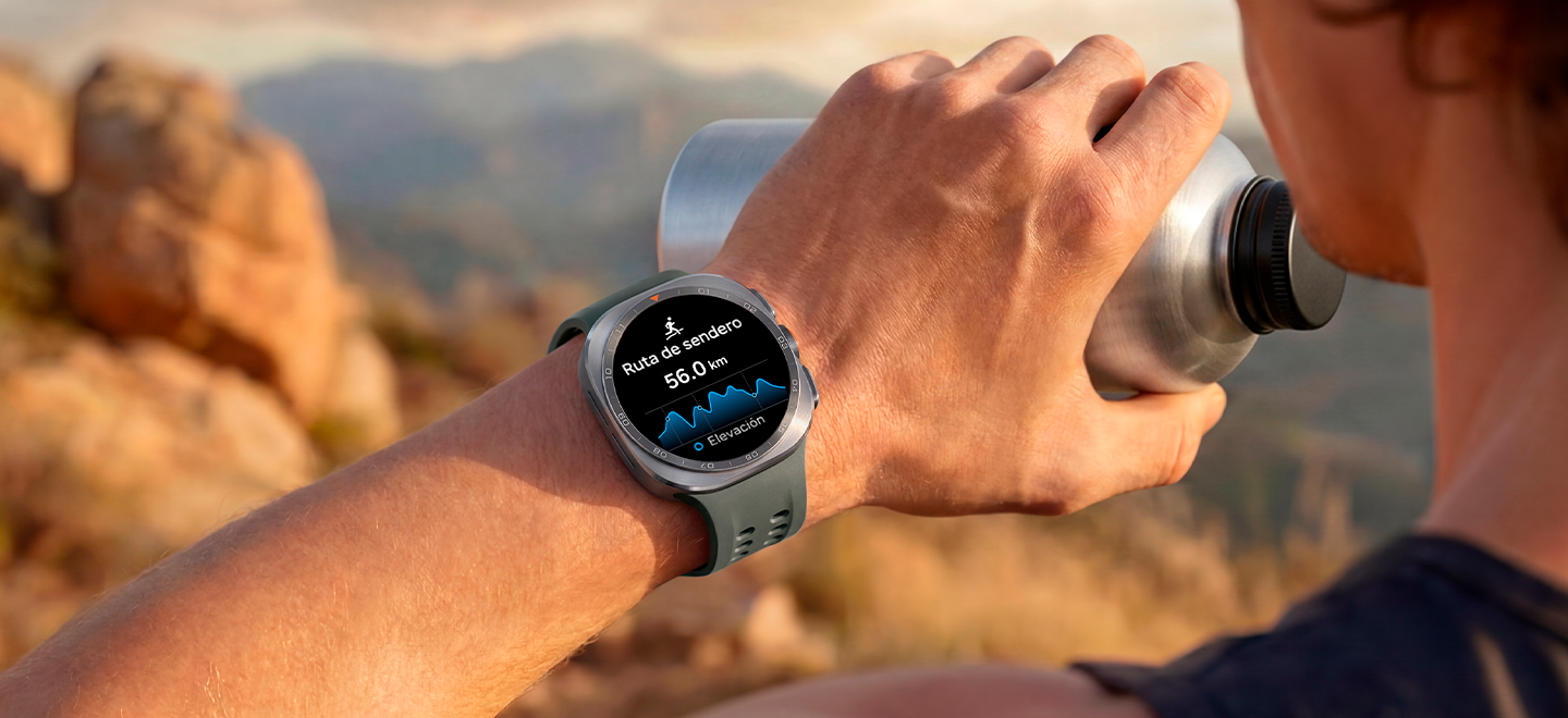 Un hombre en un sendero de montaña con terreno rocoso sostiene un vaso de agua en la mano mientras usa un Galaxy Watch Ultra2 en Titanium Silver con una Correa Marine en Olive, el cual muestra una ruta con la distancia, la mejor guía en condiciones exigentes al correr por la montaña o escalar.