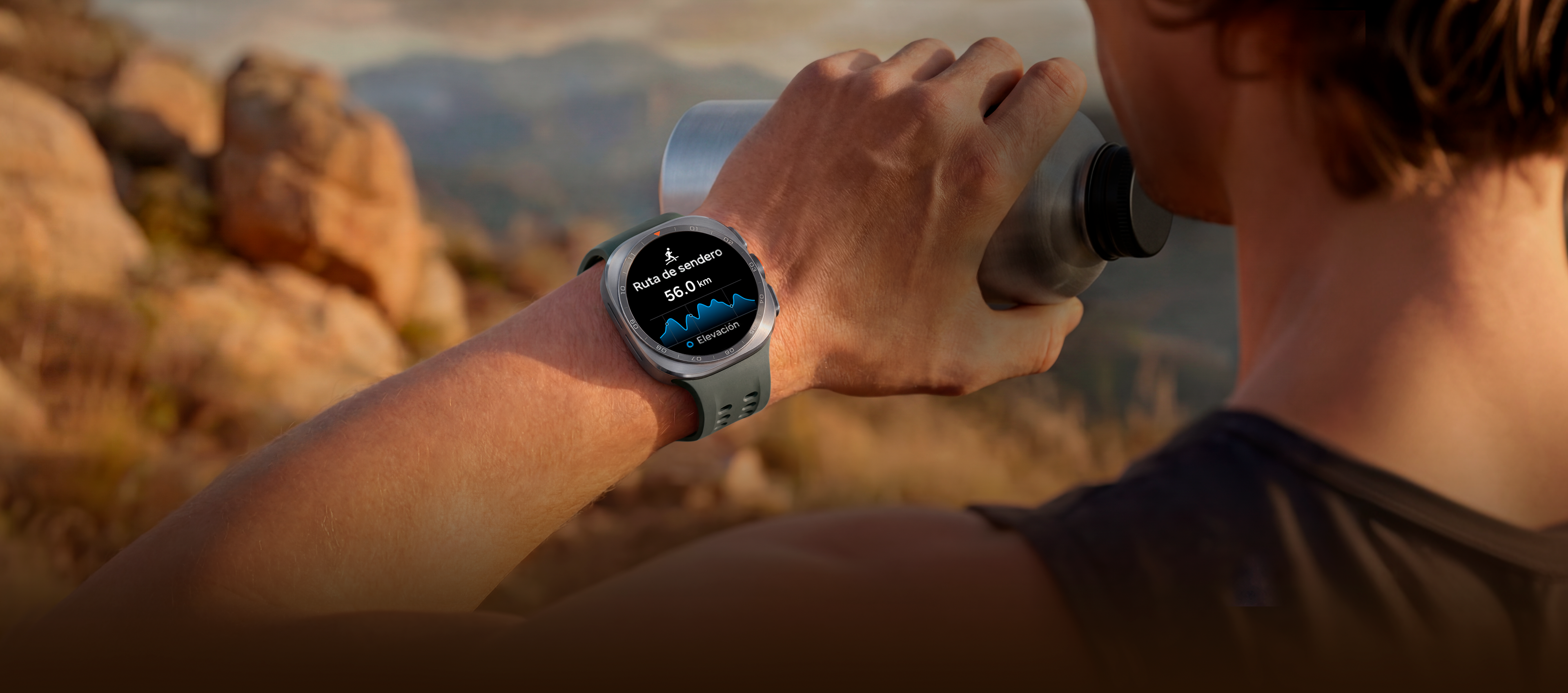 Un hombre en un sendero de montaña con terreno rocoso sostiene un vaso de agua en la mano mientras usa un Galaxy Watch Ultra2 en Titanium Silver con una Correa Marine en Olive, el cual muestra una ruta con la distancia, la mejor guía en condiciones exigentes al correr por la montaña o escalar.