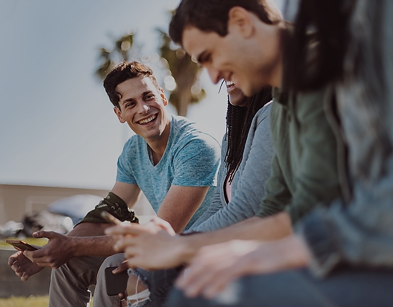 Eine Gruppe von jungen Menschen, die alle mit ihren Smartphones in der Hand zusammensitzen. Ein Mitglied der Gruppe lächelt den anderen zu.