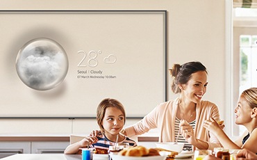 An einem Esstisch genießen 4 Personen Ihre Mahlzeiten. Im Hintergrund ist ein Super Big TV zu sehen, der an der Wand hängt. Auf ihm läuft im Ambient Modus eine Wettervorhersage.
