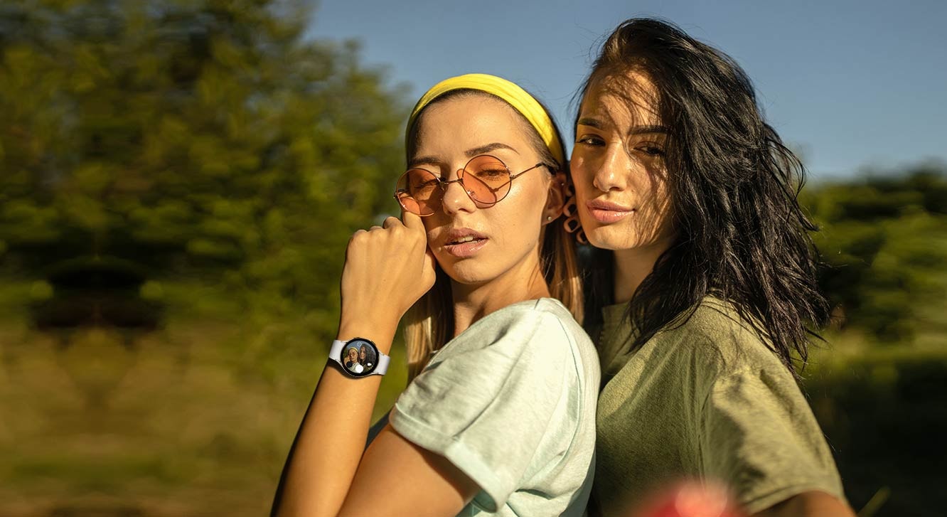 Two people pose together for a selfie, one wearing a Galaxy Watch5 on her wrist. The selfie image can be previewed in real-time shooting on the watch's display screen.