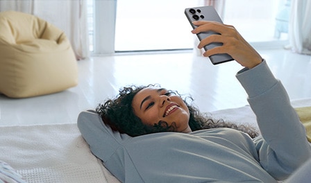 امرأة مستلقية على الأرض وهي تبتسم وتنظر إلى هاتف Galaxy S25 Ultra مع ظهور كيس حبوب أصفر خلفها.