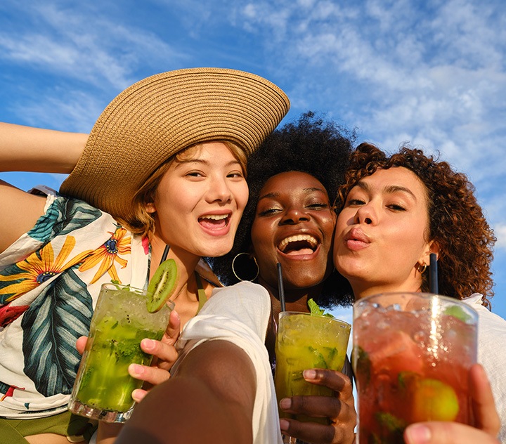 Kolme juomia pitelevää naista katsovat samaan suuntaan, ikään kuin heitä kuvattaisiin.