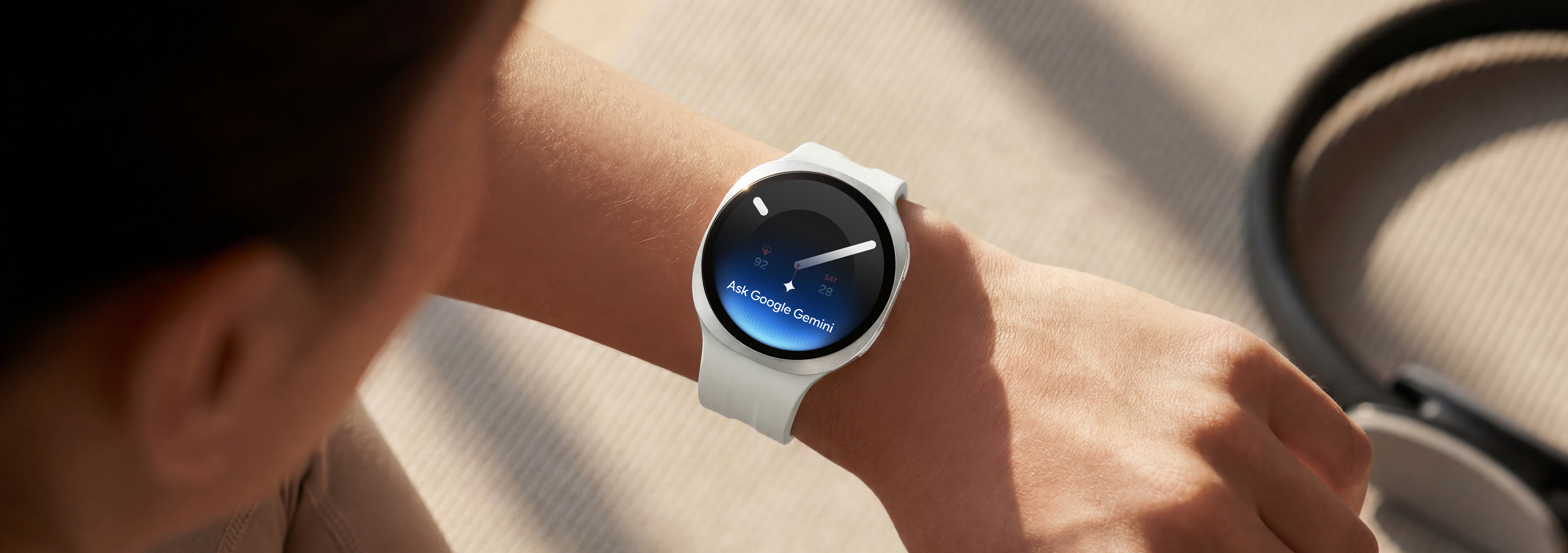 Henkilö käyttää Cream-väristä Galaxy Watch9 -kelloa, jossa on Cream-värinen urheiluranneke, ja nostaa rannettaan antaakseen kellolle äänikomennon Google Geminin avulla.