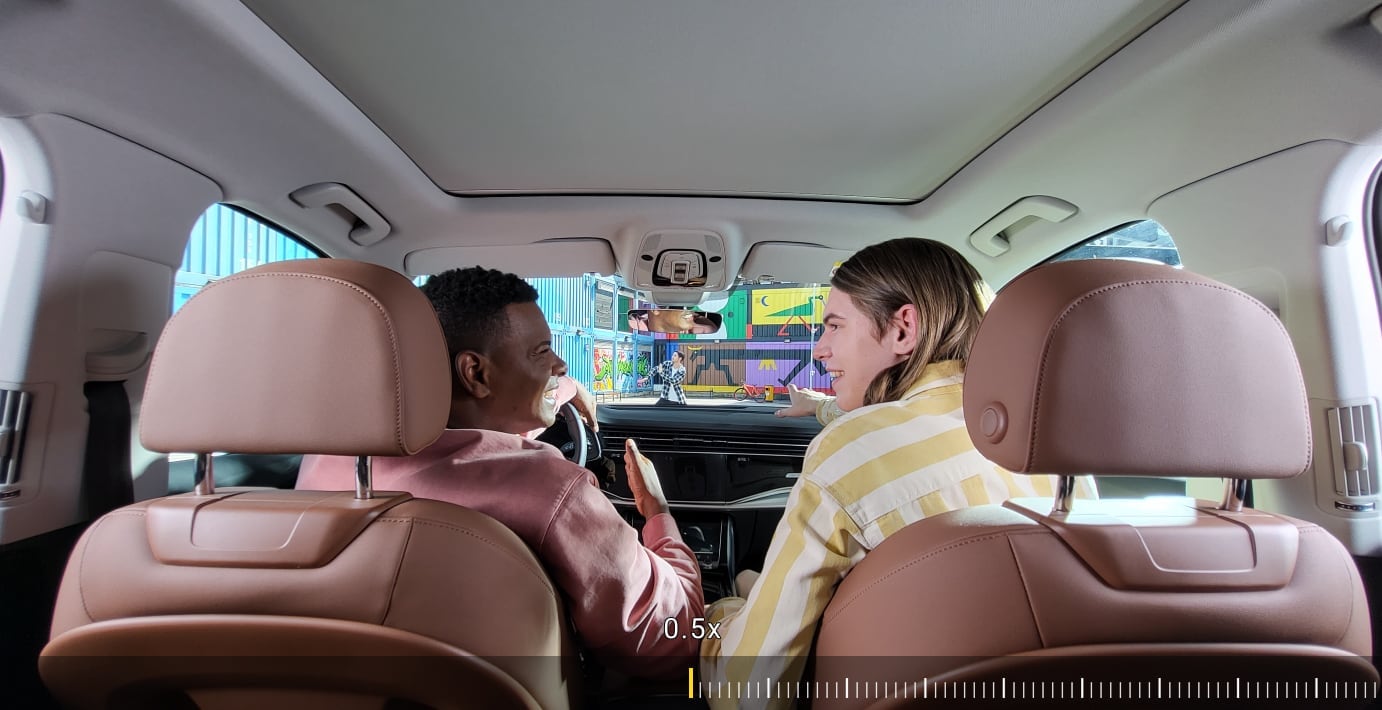 Deux amis assis à l’avant d’une voiture. Lorsque vous faites un zoom avant, vous voyez un homme passer à travers le pare-brise. Il danse en écoutant de la musique avec ses Galaxy Buds.