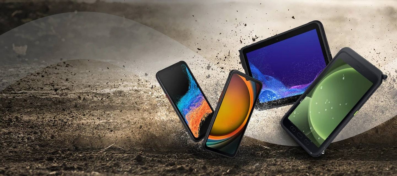 Une tablette Samsung durcie, un smartphone Samsung durci et un S Pen noir tombant sur un sol en béton gris avec des nuages de poussière gris.