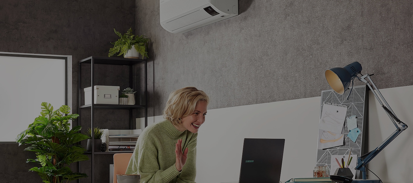 Une femme assise avec son ordinateur portable sous un climatiseur accroché au mur et agitant les mains.