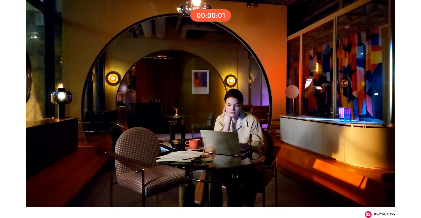 शीर्षस्थानी टाइमरसह एक व्हिडिओ रेकॉर्डिंग करीत आहे. एक व्यक्ती, त्याच्या लॅपटॉपसह रात्री गडद कॅफेमध्ये बसलेला. ती वर पाहते आणि हसते. तळाशी उजवीकडे एक कॅमेरा चिन्ह आहे जो त्याच्या पुढे 