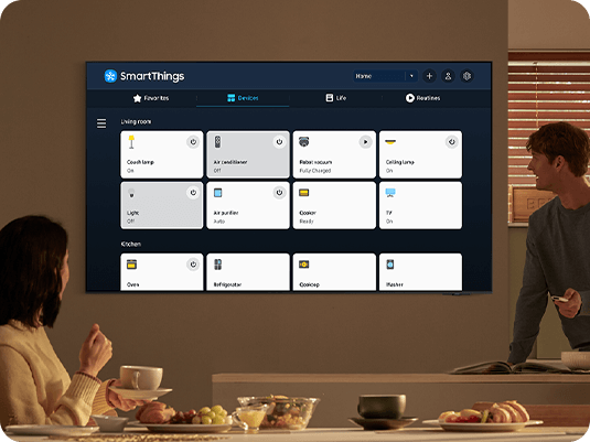 Two people are looking at the 2024 Samsung OLED TV screen and controlling their devices with the SmartThings app that makes their house smart.