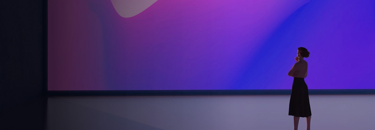 Pohled zezadu na ženu stojící na jevišti a hledící na velkou obrazovku, na které jsou zobrazeny jasně modré a fialové barvy.