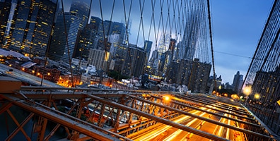 An image of a bridge made of steel with a bent angle and a city background behind it.