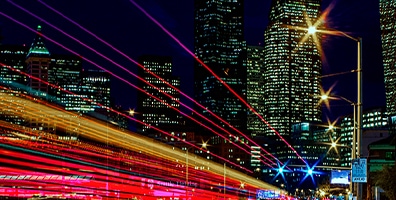 An illustrative image of a night view of the city with high-rise buildings in the back, and Red, yellow, and white thin lines extend from front to back along the driveway, and these lines represent a 5G network.