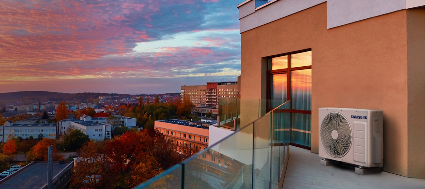 A Samsung Free Joint Multi outdoor unit sits on a balcony overlooking a cityscape with colorful autumn trees. Clouds drift by as the sun sets, casting a warm glow.