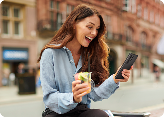 A woman is sitting in an outdoor location. She smiles as she looks down at her phone, viewing her pet at home.