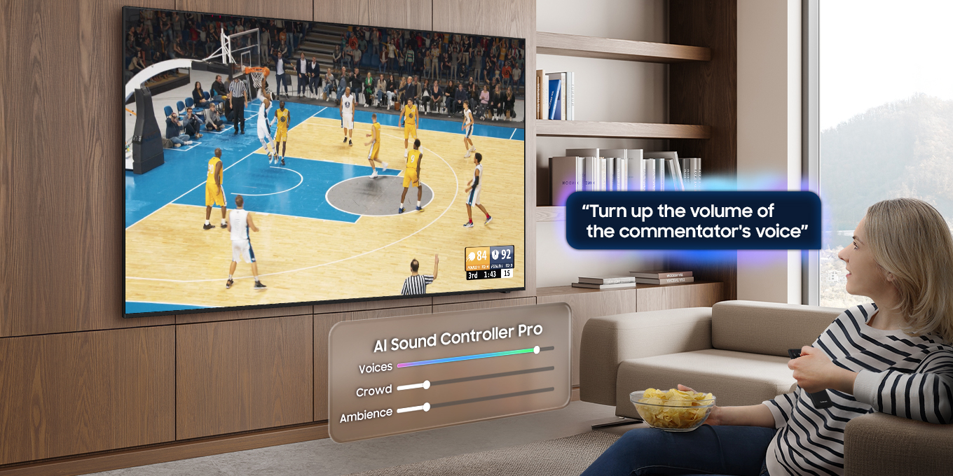 سيدة تشاهد مباراة كرة سلة على تلفزيون Samsung TV وتتحدث إلى جهاز تحكم عن بُعد. تقول ”ارفع مستوى صوت المعلّق”. وأسفل التلفزيون، تعرض ميزة AI Sound Controller Pro مستوى الصوت للحوار والجمهور والأجواء بشكل منفصل، مع رفع مستوى صوت الحوار.
