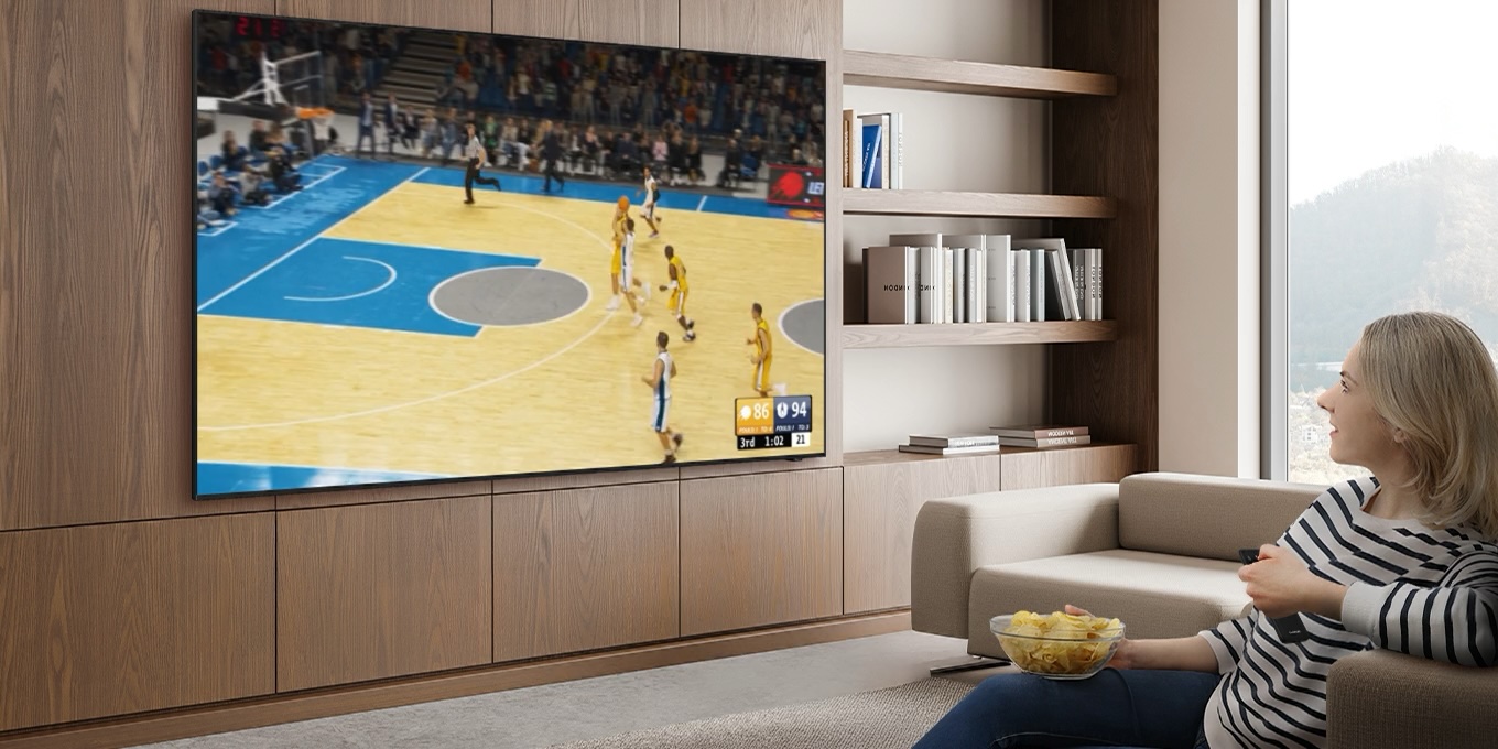 سيدة تشاهد مباراة كرة سلة على تلفزيون Samsung TV. ثم تتحدث إلى جهاز التحكم عن بُعد قائلة: ”ارفع مستوى صوت المعلّق”. أسفل التلفزيون، تعرض ميزة AI Sound Controller Pro مستويات الصوت لثلاثة مسارات يتم تعديلها وفقاً لذلك، مع رفع مستوى أصوات الحوار وخفض مستوى صوت الجماهير والأجواء.