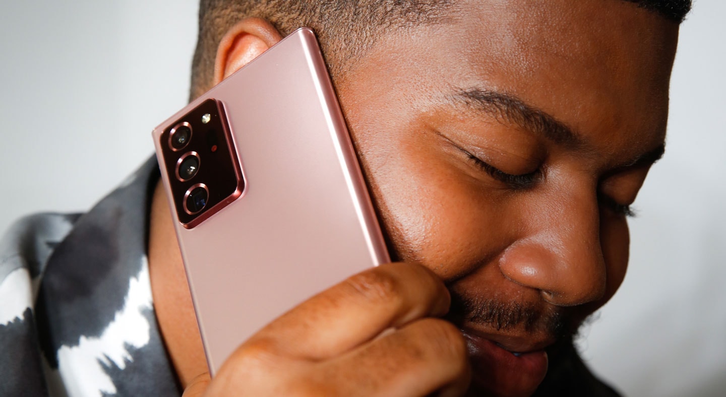 Khalid hält ein Galaxy&nbsp;Note20 in Mystic&nbsp;Bronze in einer Hand und den dazugehörigen S&nbsp;Pen in der anderen und lacht. Er sitzt auf einer beigefarbenen Couch und trägt ein schwarz-weißes Batik-T-Shirt.