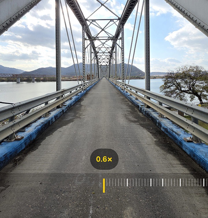 Camera feature onscreen. View of a bridge can be seen. The zoom goes up from 0.6x to 100.0x and shows the backside of a person that is far away. Zoom goes down to 10.0x and shows the person with the bridge. On the bottom right is a camera logo with text Captured by Galaxy S25 Ultra hashtag with Galaxy.