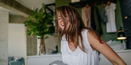 A woman sits up in bed in her room, laughing with her eyes closed as her hair covers half of her face. She is wearing a white sleeveless tank top.