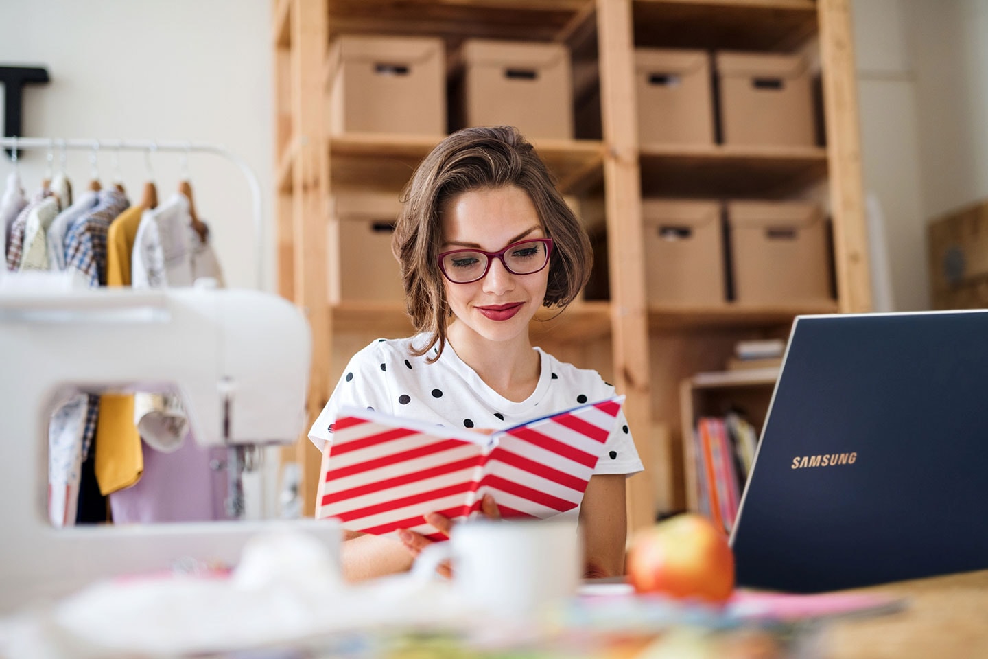 A woman checks her notebook while working in her studio on a Samsung laptop. A rack of clothing and a shelf of boxes are behind her.