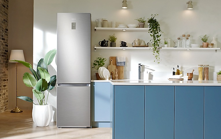 Samsung hladnjak stoji između zelene biljke i kadetskih plavih sudopera u modernoj minimalističkoj kuhinji. Police su naslagane nekim tanjurima i ukrašene malim biljkama.