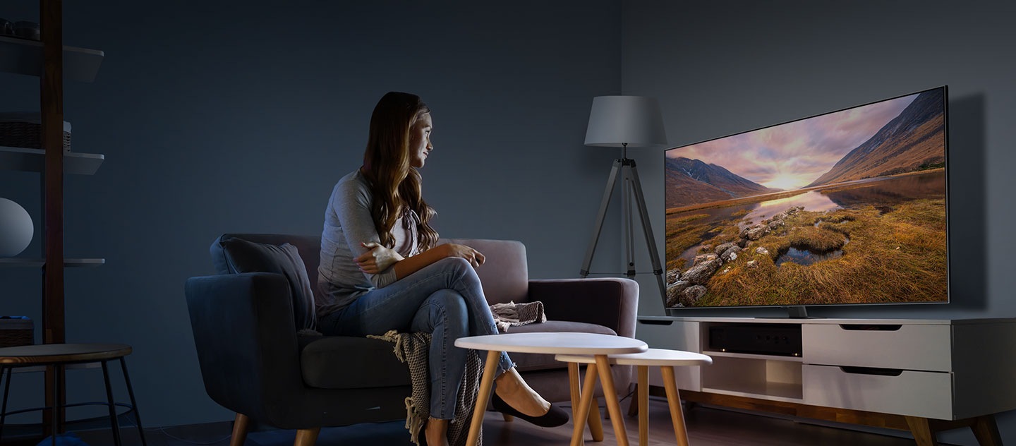 A woman sitting down on a sofa in front of a Samsung TV, viewing a majestic landscape image on the screen cast from her Galaxy smartphone.