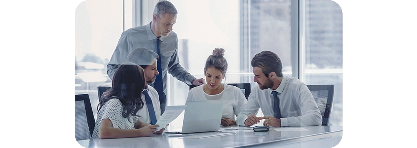 Five people in a meeting room looking at something on a laptop and taking notes
