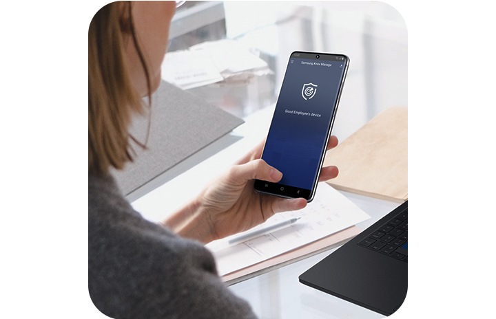 A woman sitting at a desk holding her Galaxy S20 Ultra, which has the Knox Manage interface onscreen