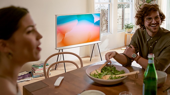 A man and a woman are enjoying a meal at the kitchen table, laughing and talking with a third person who is unseen. A Cloud White model of The Serif is showing colourful image using Ambient mode in the background.