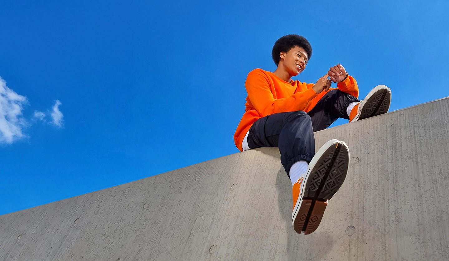 A young man in an orange t-shirt sits on top of a wall with one leg hanging down while tapping on his Galaxy Watch to take his selfie from a lower angle, taken with a Galaxy Z Flip3 on Flex mode.