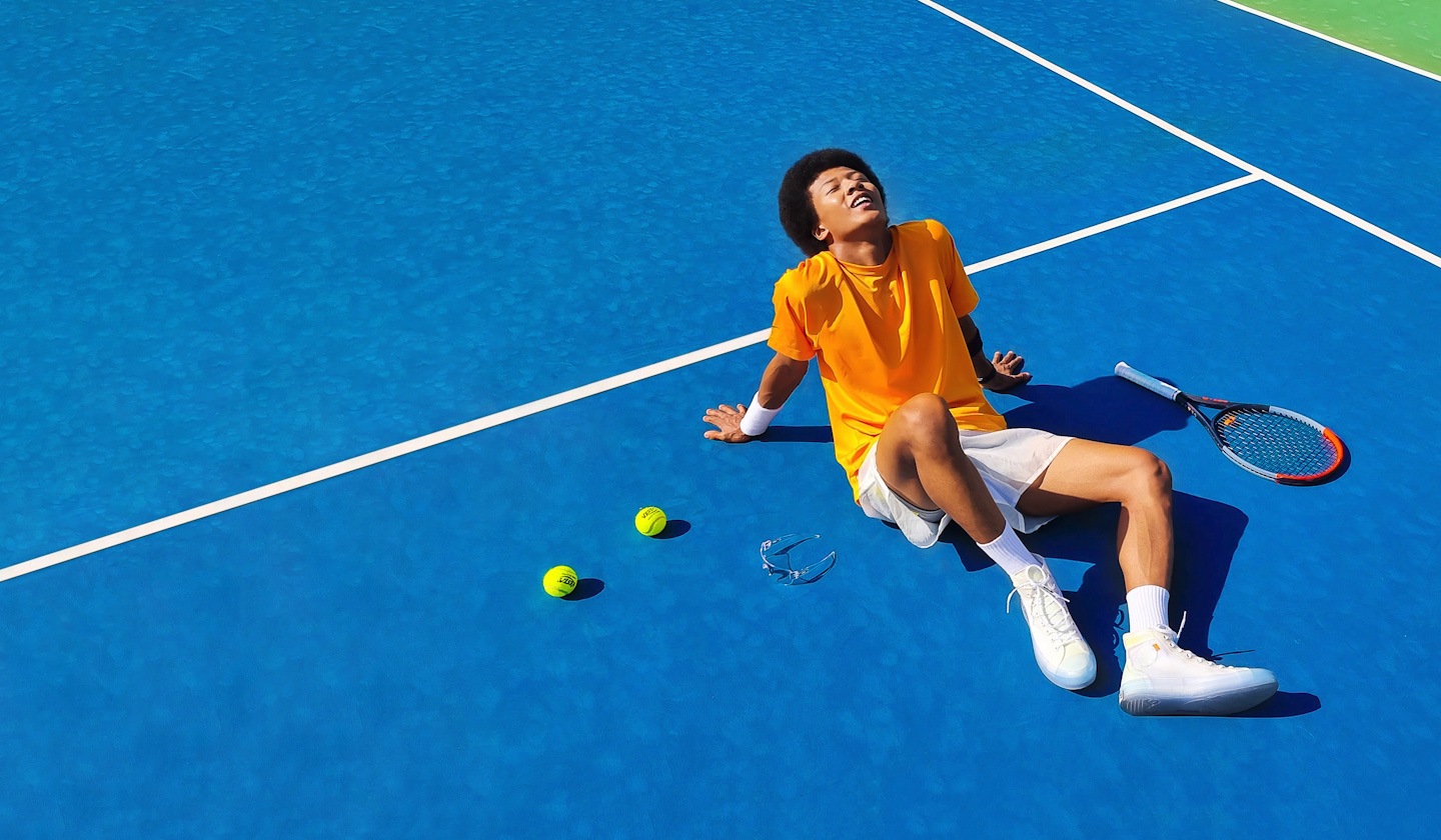 A young man in an orange t-shirt is sitting down on a sun-lit, bright blue tennis court, seemingly enjoying the sunlight. On the lower left corner, an intruding shadow of a lighting post nearby the tennis court is promptly erased.