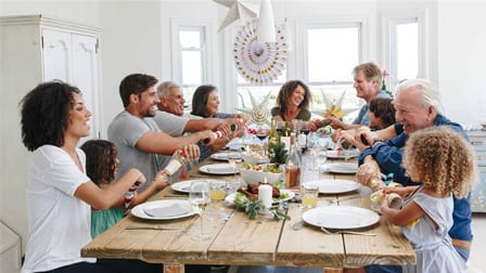 A smiling family sits around a dining room table getting ready to pull Christmas crackers