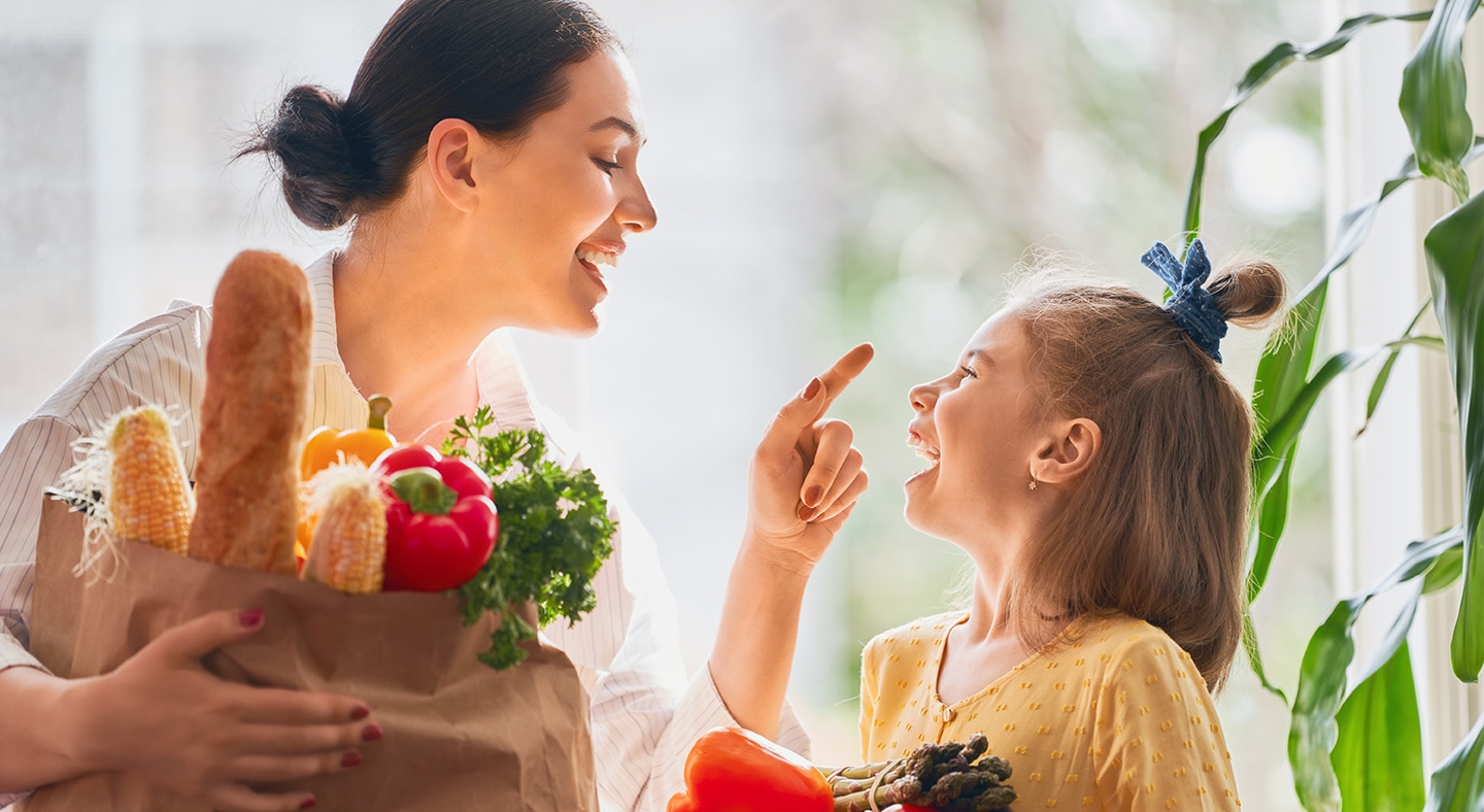 Mother and her daughter looking at each other with big smiles, holding grocery shopping bag filled with vegetables.