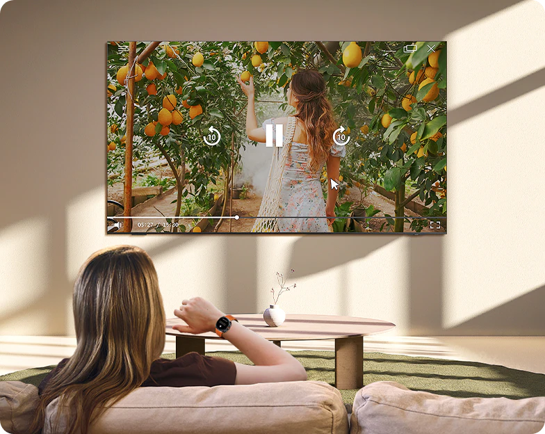 A woman controls a TV across the room with her Galaxy Watch. She gestures to press pause on a scene of a girl picking oranges.