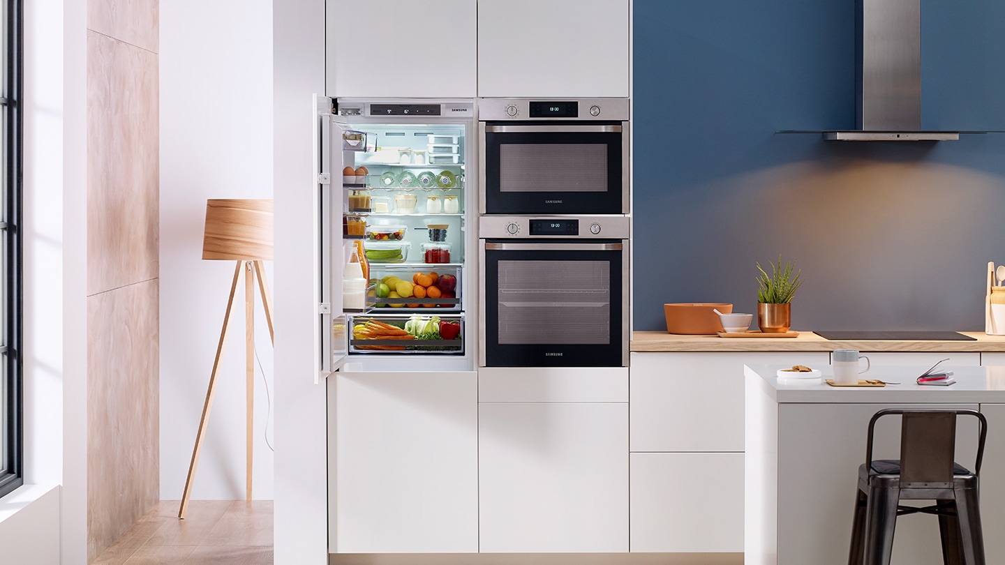 Un intero set di elettrodomestici da incasso Samsung come frigoriferi, forni e forni compatti installati in una cucina bianca premium. Mentre la porta del frigorifero è aperta…