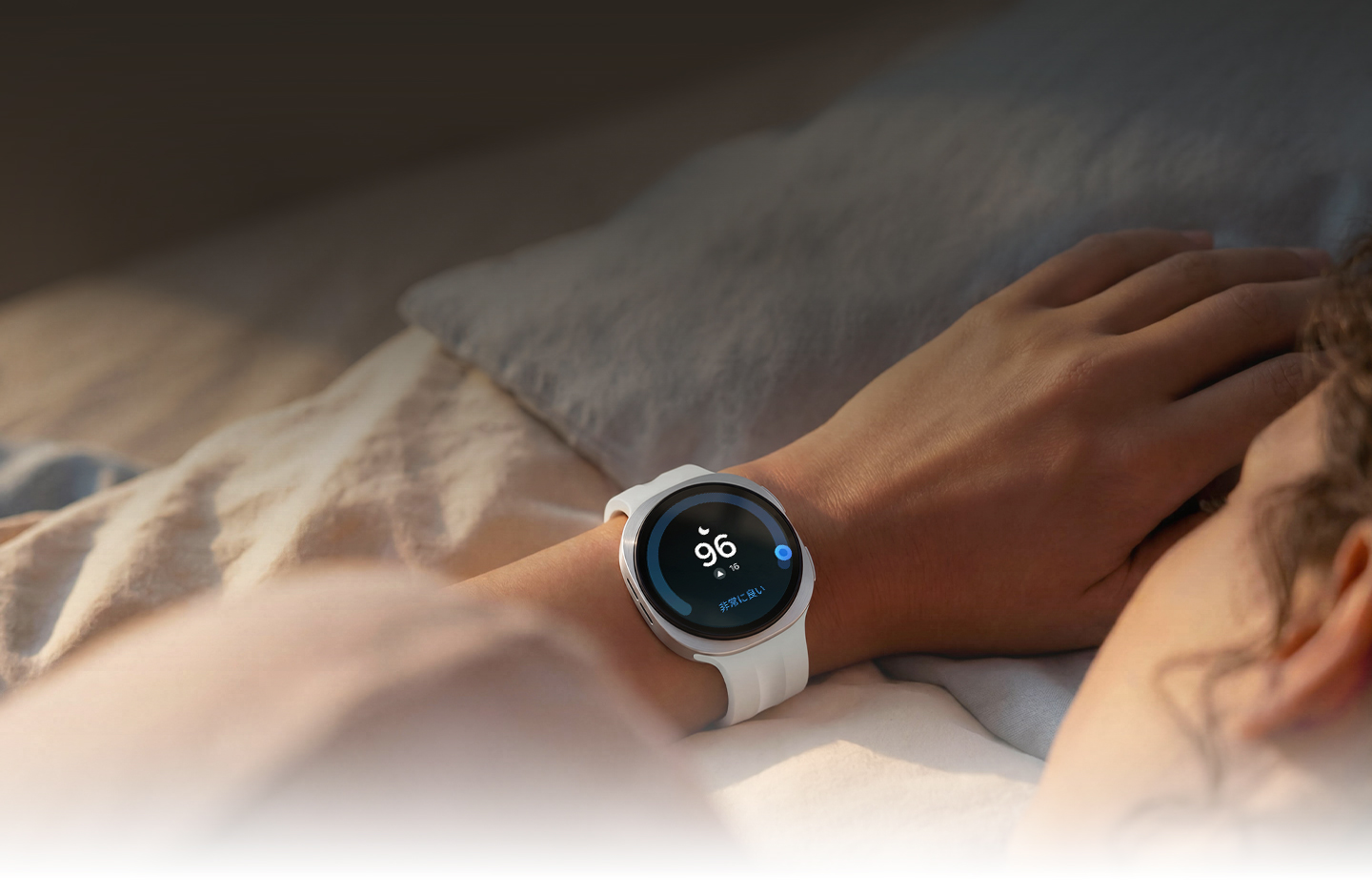 Galaxy Watchを手首に装着した状態でベッドに横たわっている人がいる。睡眠マークと96という数字が画面に表示されており、ウォッチが睡眠中の呼吸の乱れを繰り返しトラッキングしていることを示している。睡眠トラッキング機能のGUIがGalaxy Watch9の近くに表示されている。睡眠ステージのデータ、睡眠時間の一貫性、エナジースコアが表示されている。